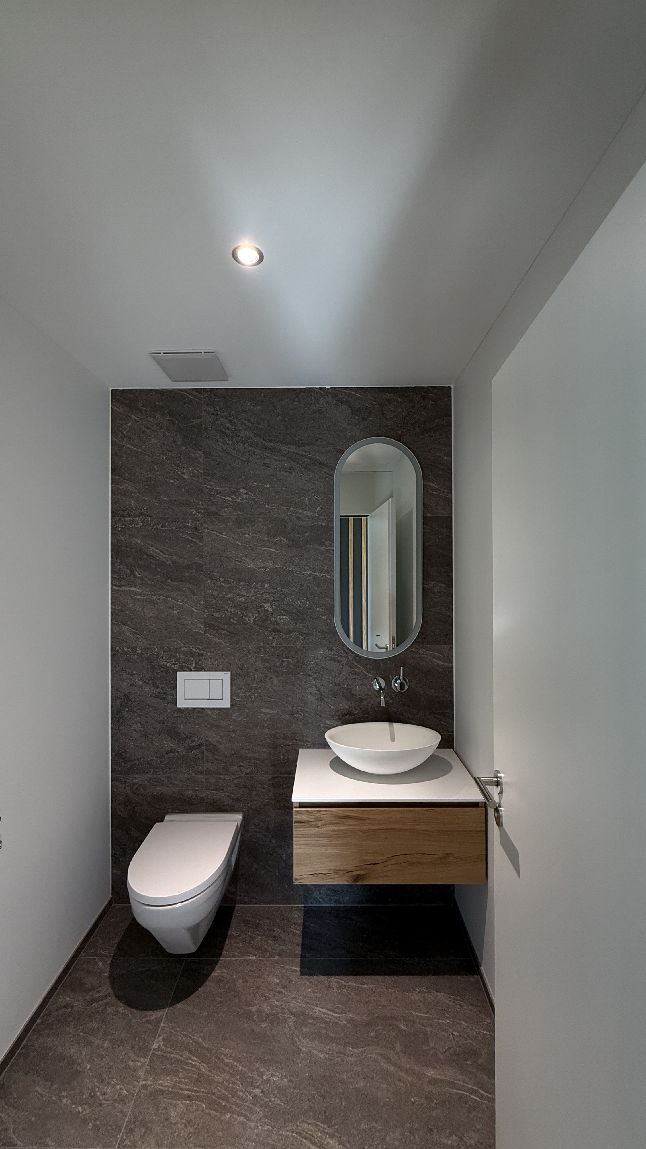 Rothusweg — Gäste-WC mit dunklen Natursteinfliesen, ovalem Spiegel und schwebendem Eichenwaschtisch